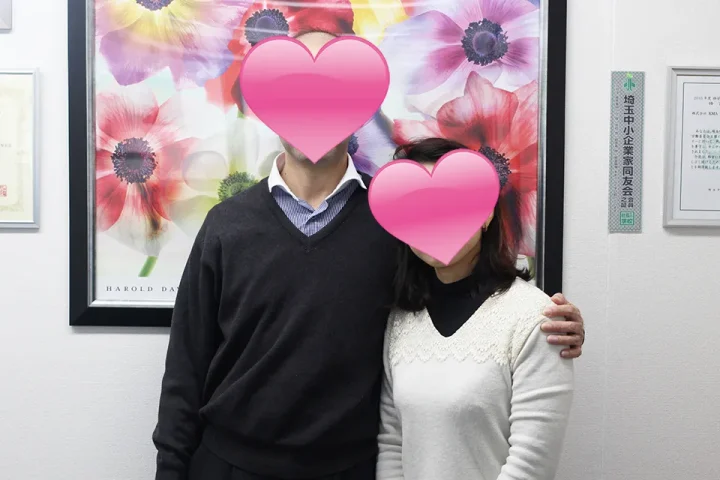 相談室で肩を寄せ合う40代バツイチ女医と公務員男性の成婚ツーショット写真。花の絵の額の前で幸せそうに微笑むカップル。