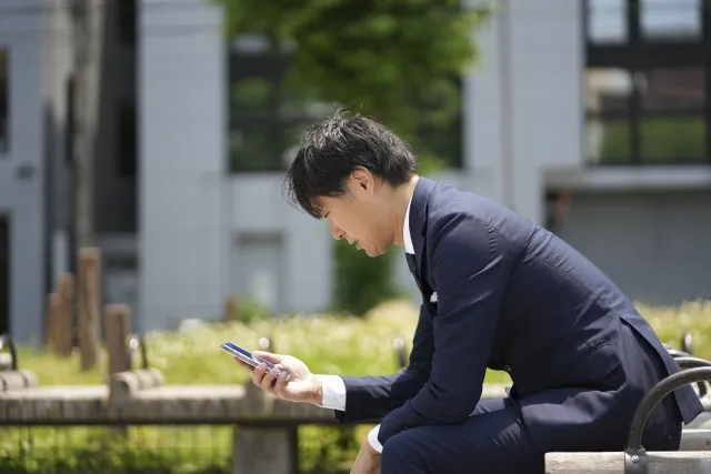 公園のベンチに座り、スマートフォンを見ながら仮交際終了の連絡に戸惑う婚活中の男性