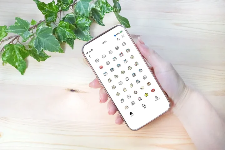 婚活女性が白い木目調のテーブルでスマホを持ち、LINEに「ちいかわ絵文字」を並べて操作する手元。テーブルには観葉植物が置かれ、婚活LINEで気持ちを柔らかく伝える場面を表現。