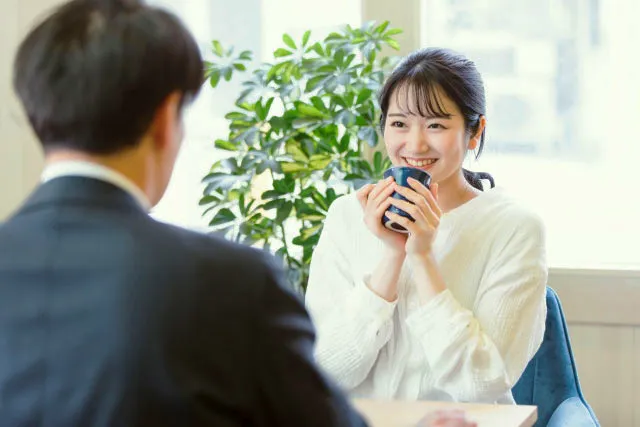 落ち着いた雰囲気のカフェで、笑顔でコーヒーカップを持ち、男性と会話する30代独身女性。医師とのお見合いをイメージしたシーン。