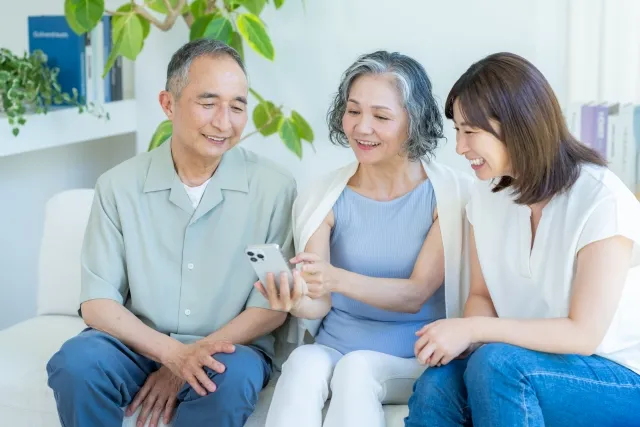 30代独身女性が実家のリビングで父母と並んでスマホをのぞき込み、笑顔で過ごす様子。家族との穏やかな時間を楽しむ実家暮らしの一コマ。