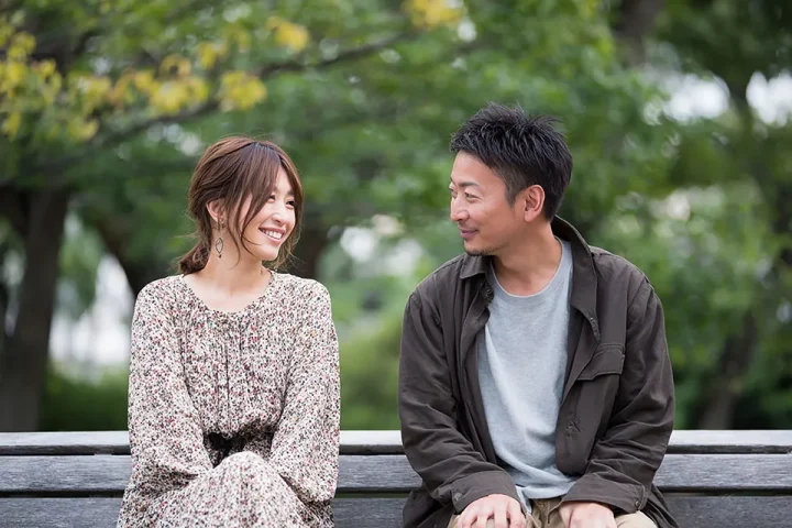 公園のベンチに座って笑顔で会話する30代男女。穏やかな初秋の風景の中、自然な雰囲気で婚活について話している様子