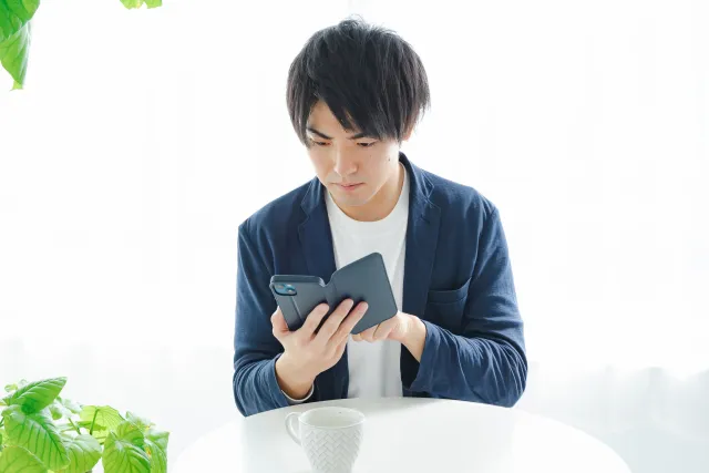 明るい室内で真剣な表情でスマートフォンを見つめる男性。観葉植物とコーヒーカップが置かれ、婚活デートの会話に悩む様子を表した写真