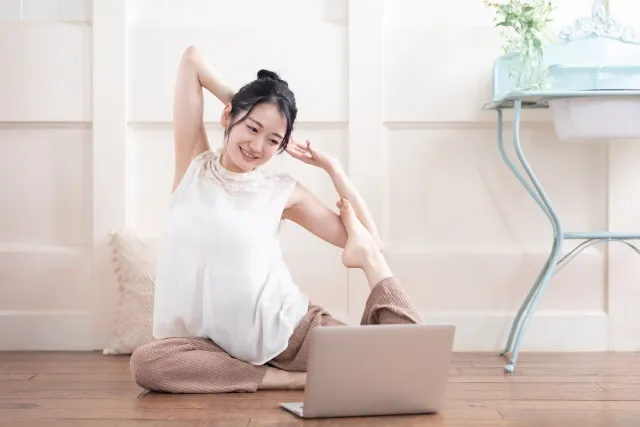 明るい室内でオンラインヨガをする女性。パソコンの画面をにこやかな表情で見ながら「鳩のポーズ」を決める様子