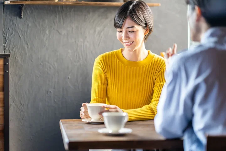 カフェでのお見合い風景。女性がコーヒーカップに手を添えながら、男性の話に耳を傾け、はにかんだ表情でそっと視線を外している様子。初対面の緊張感と心の距離が少しずつ縮まる瞬間を描いた温かい場面