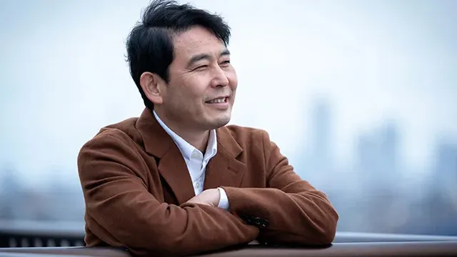 都心のビル屋上で腕を組み、はにかんだ笑顔を見せる50代男性