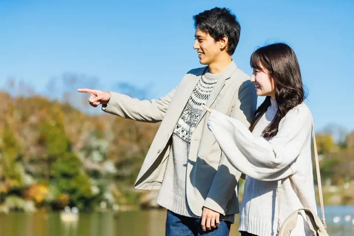 秋の水辺を仲よく歩く男女が、指さす方向を笑顔で見つめる姿。将来への希望と心のつながりを感じさせる一枚