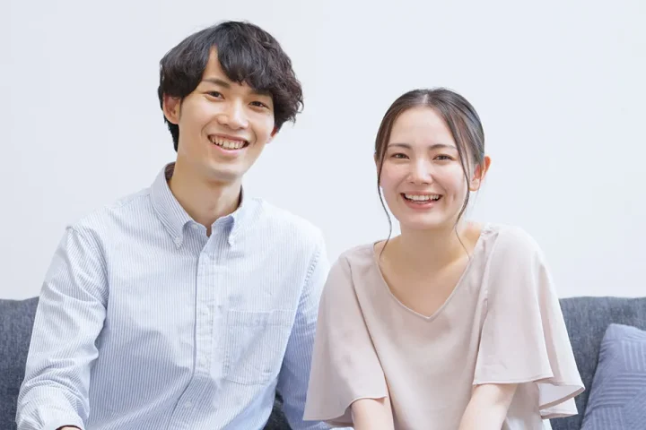 ソファーに肩を並べて座る、笑顔の若いカップル。穏やかな雰囲気の中で、信頼と安心感が伝わる一枚