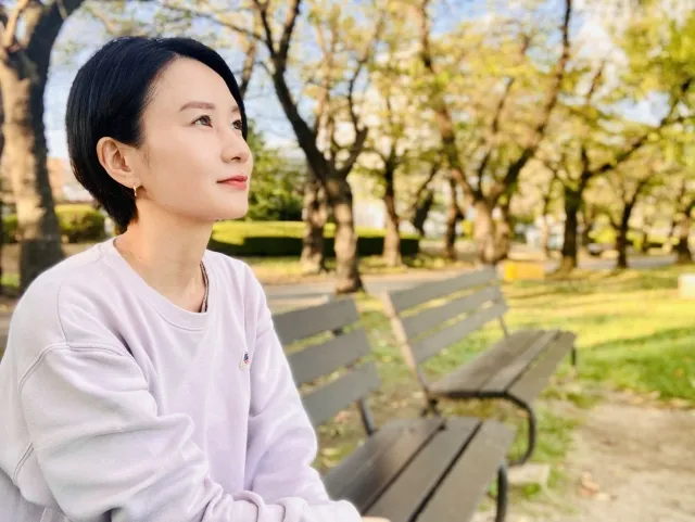 秋の公園のベンチで遠くを見つめながら考え事をする女性の横顔。柔らかい日差しが心の揺れを包み込むように差し込んでいる