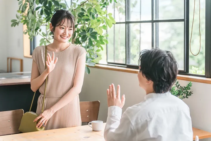 カフェで別れ際、笑顔で手を振る男女。男性は席に座り、女性は立ち上がって優しく手を振りながら去っていく