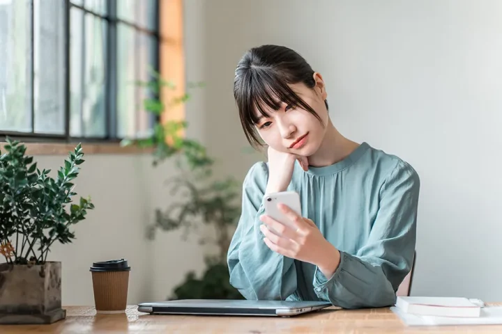 仮交際中の女性がカフェでスマホを見ながら悩んでいる様子。誰と真剣交際に進むか迷っている表情