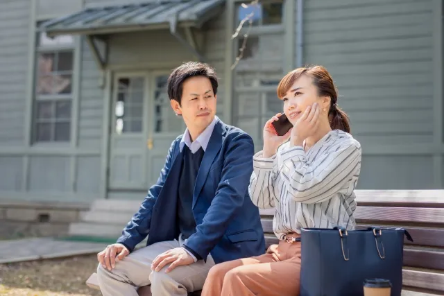 ベンチに並んで座る男女。女性は斜め上の空を見上げながら笑顔で携帯電話を使い、男性は少し不安そうな表情で彼女を見つめている婚活シーン