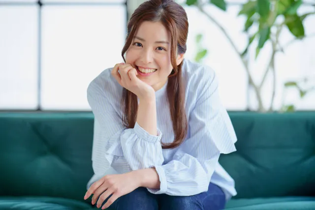明るい室内で頬杖をつきながら柔らかく微笑む女性の写真。愛嬌のある自然な笑顔が伝わるイメージ