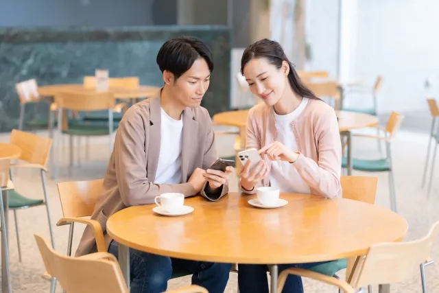 カフェで微笑みながらスマホを見せ合うカップル。自然体で会話を楽しむ様子
