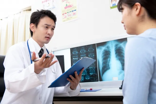 バインダーを持った男性医師が、女性患者の話に丁寧に耳を傾けている様子。信頼感と誠実さを感じさせるシーン。