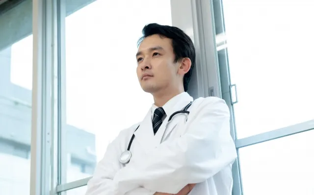 病院の廊下で窓にもたれ、腕を組んで思索にふける男性医師。多忙さと責任の重さを象徴するシーン。