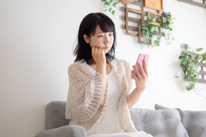 スマートフォンを不安そうに見つめる女性がソファーに座っている様子。婚活アプリに悩む気持ちを表現した写真。