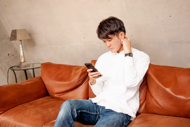 自宅のソファでスマホを見ながら悩む男性。アプリ婚活の迷いや不安を抱える様子を表現したイメージ写真。