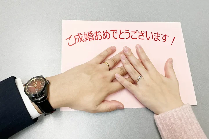 成婚カップルが『ご成婚おめでとうございます』のペーパーの上で指輪をはめた手を重ねる写真｜埼玉県さいたま市の結婚相談所KMA