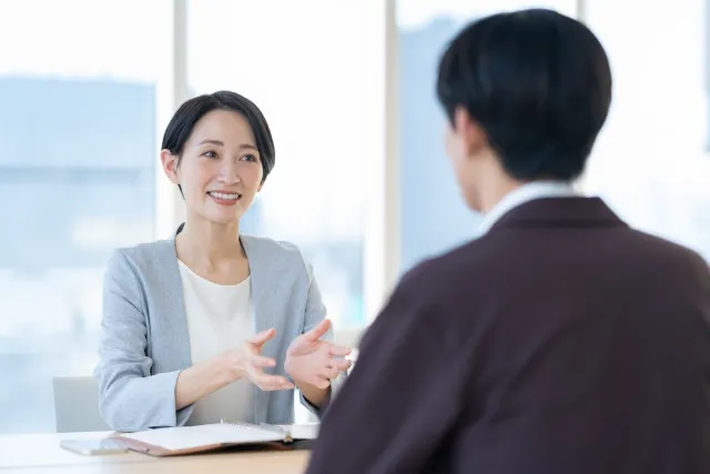 明るい室内でカウンセラーが資料ファイルを見せながら、若い男性に丁寧に説明している無料相談の様子。テーブルにはスマートフォンと資料が置かれている。