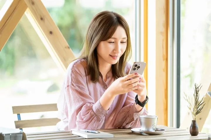明るいカフェでスマホを見ながら微笑む女性。テーブルには観葉植物と手帳とコーヒーが置かれ、気軽に婚活を始める雰囲気を表現した写真。