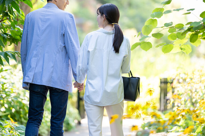 公園の緑道を寄り添って歩く40代カップルの後ろ姿。自然体で会話を楽しむ様子が伝わる写真