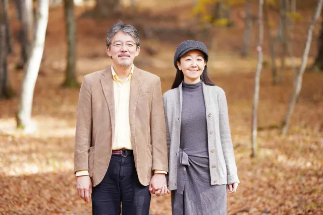 晩秋の自然の中で、手をつなぎながら穏やかな笑顔で並んで立つシニアカップルのひざ上ショット