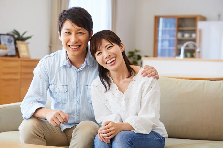 ソファーに座り、男性が女性の肩を優しく引き寄せながら笑顔でカメラを見つめるカップル。信頼と親しみが伝わる一枚