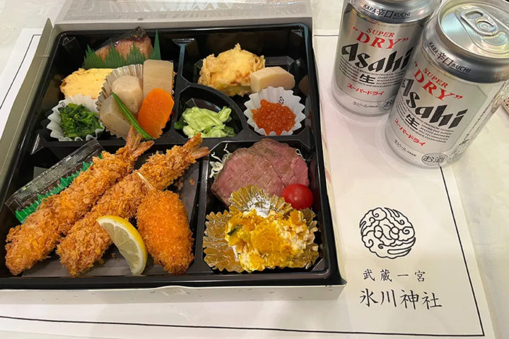 節分祭の食事会で振る舞われた会席弁当とビール。敷紙には神紋と武蔵一宮氷川神社の文字が印刷されている様子 